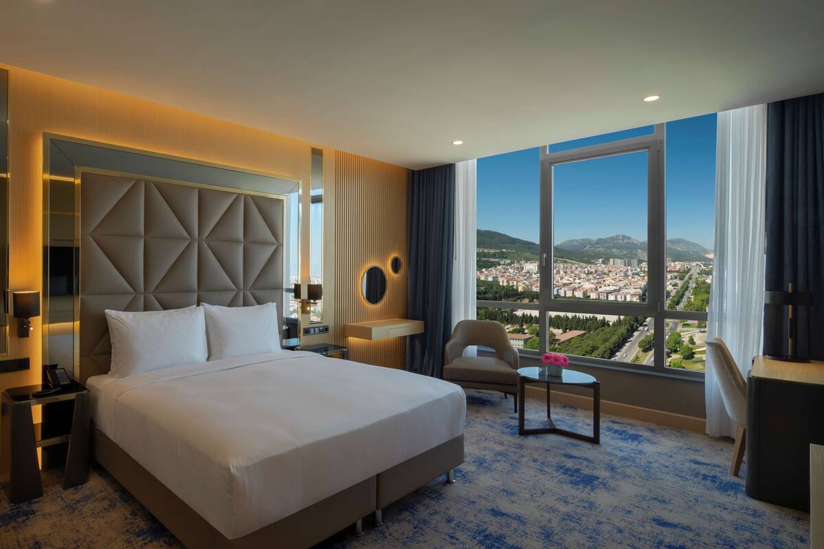 A hotel room with a large bed, a chair, a small table, a TV, and a view of a cityscape through large windows.