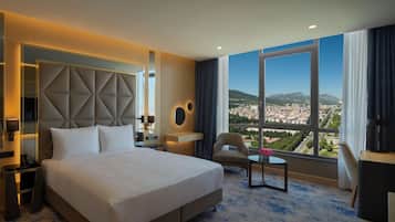 A hotel room with a large bed, a chair, a small table, a TV, and a view of a cityscape through large windows.