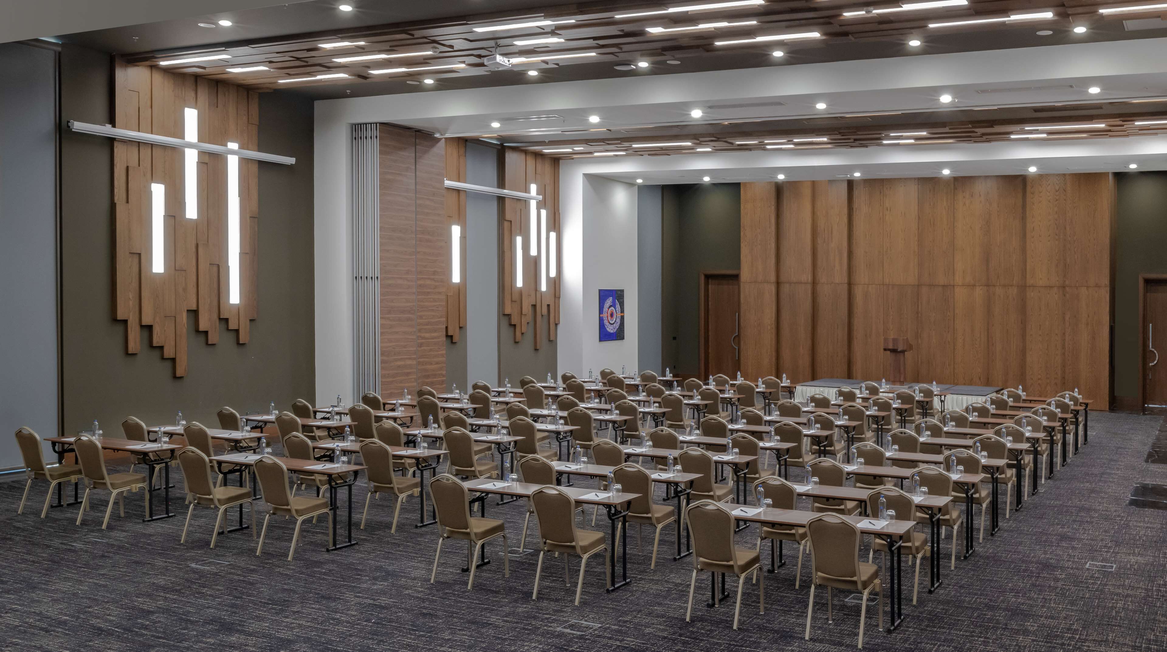 Un salón de conferencias con filas de mesas y sillas, paredes de paneles de madera e iluminación empotrada.