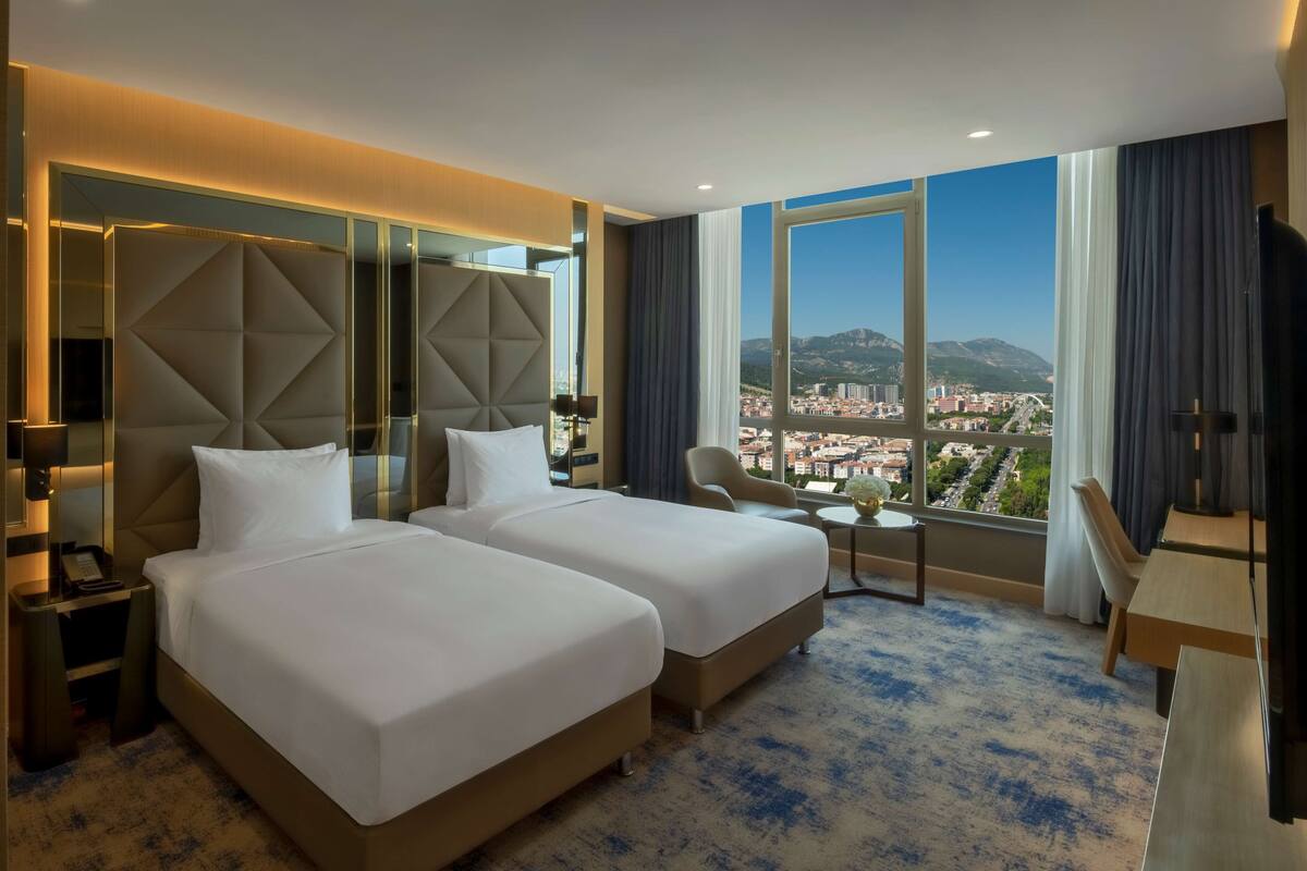 A hotel room with two beds, a small table, a chair, and a large window offering a city view.