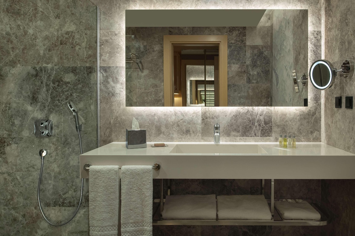 Un baño moderno con un espejo grande, una cabina de ducha de vidrio y un lavabo blanco con dos botellas de champú.