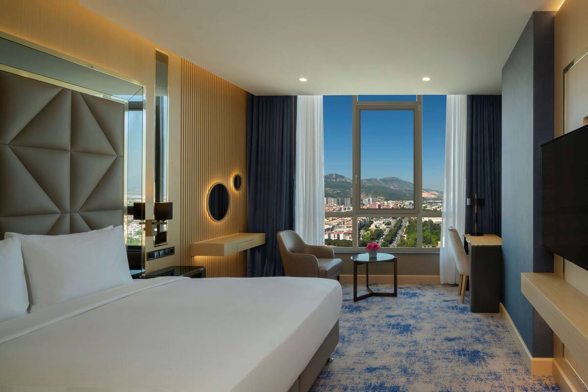 A hotel room with a large bed, a desk, a chair, a TV, and a view of a cityscape through a large window.