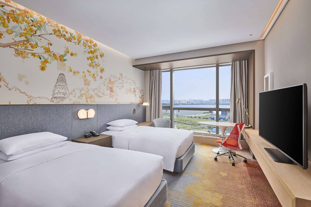 Ein Hotelzimmer mit zwei Betten, einem großen Fernseher und Blick auf eine Stadtlandschaft durch große Fenster.