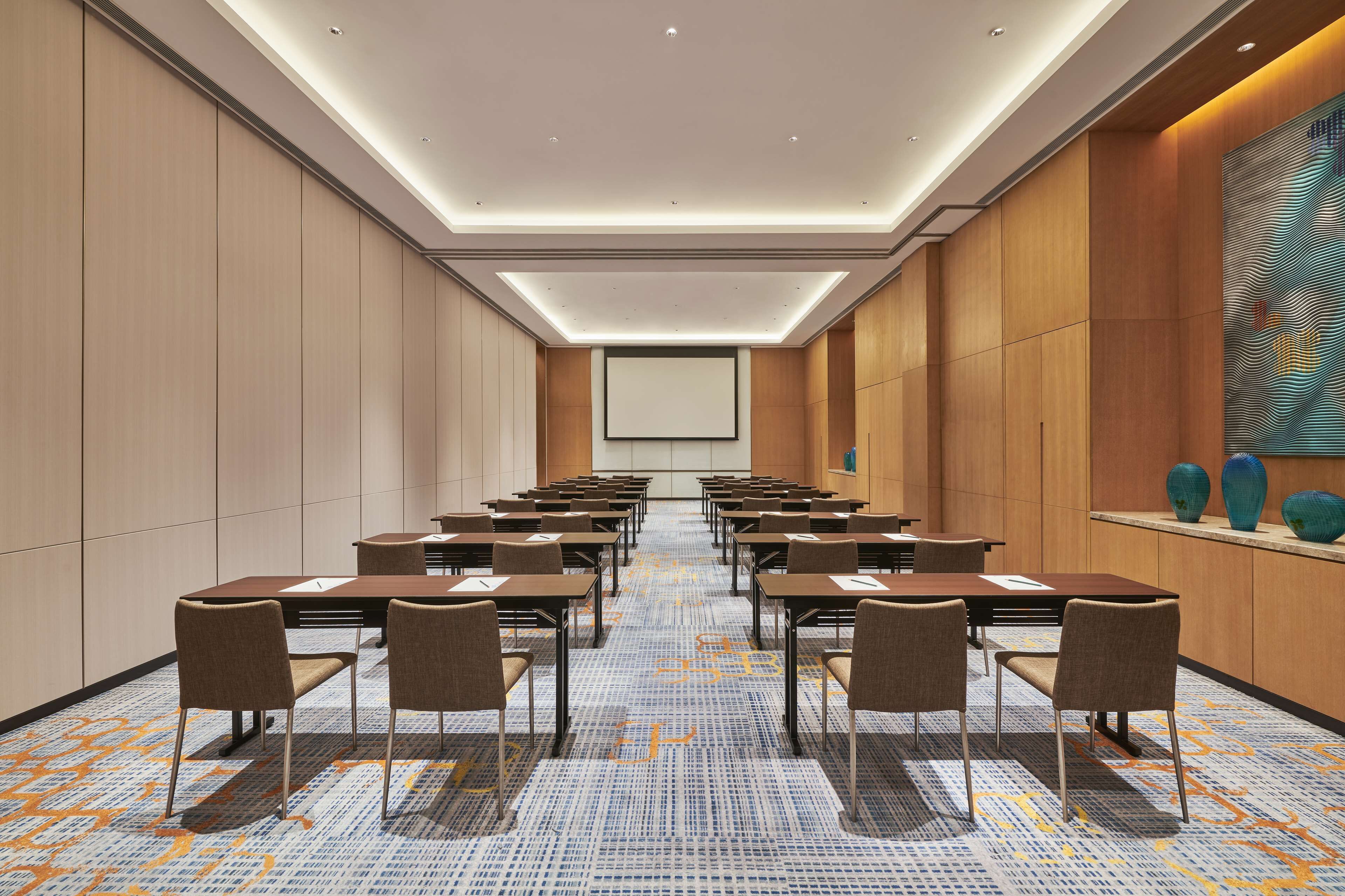 A conference room with rows of tables and chairs, a large screen at the front, and a patterned carpet.