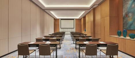 A conference room with rows of tables and chairs, a large screen at the front, and a patterned carpet.