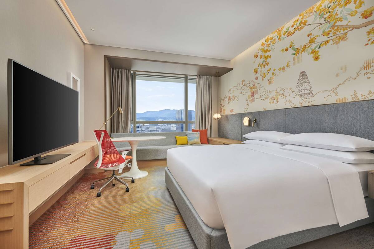 Ein Hotelzimmer mit Bett, Schreibtisch mit Fernseher, Stuhl, Fenster mit Stadtblick und einer Wanddekoration.