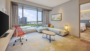 Une chambre d’hôtel moderne dotée d’une grande fenêtre, d’un canapé, d’une table basse, d’un téléviseur et d’un bureau avec une chaise.