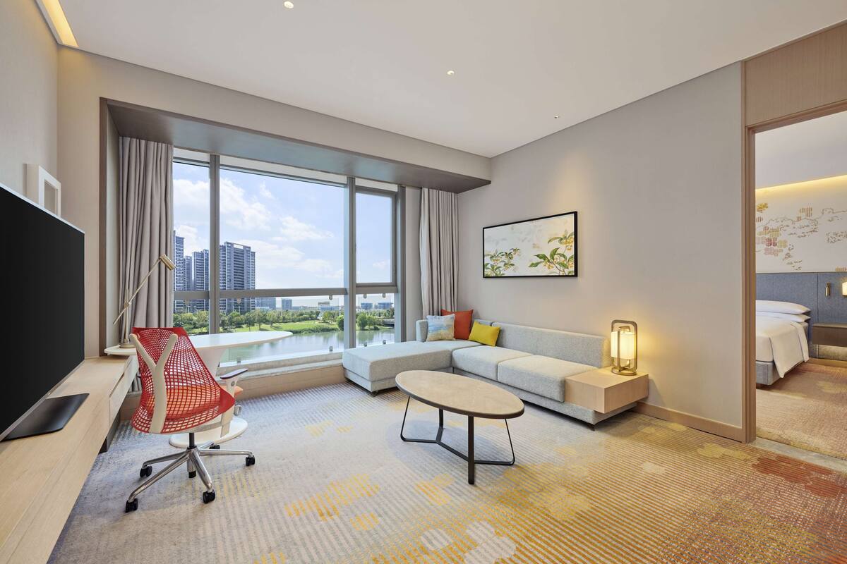 Ein modernes Hotelzimmer mit großem Fenster, einer Couch, einem Couchtisch, einem Fernseher und einem Schreibtisch mit Stuhl.
