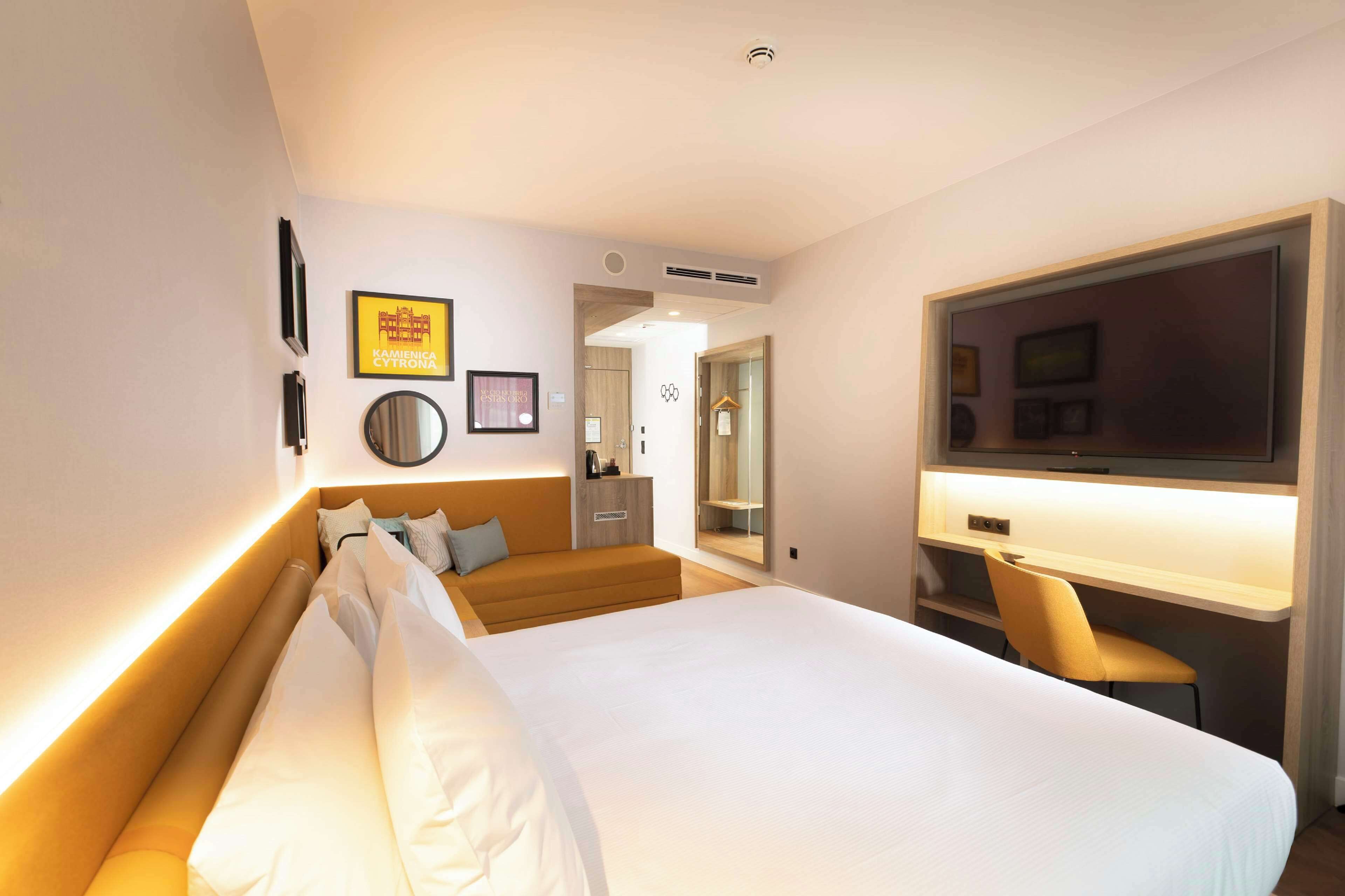 Quarto de hotel com uma cama grande, uma escrivaninha com cadeira, uma televisão e um espelho.