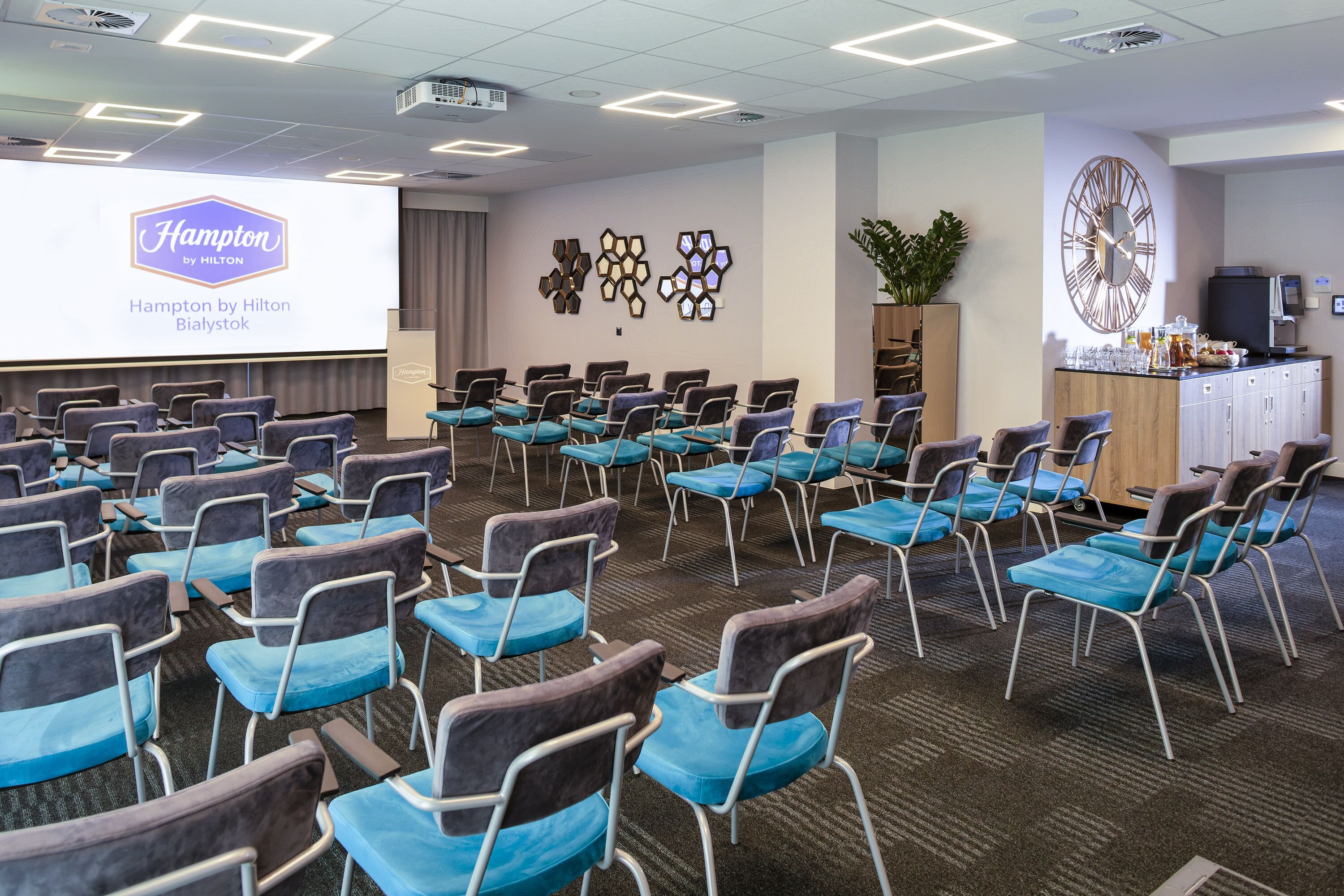 Un salón de conferencias con filas de sillas, una pantalla grande mostrando 'Hampton by Hilton Bialystok', un reloj de pared y una zona de bar.