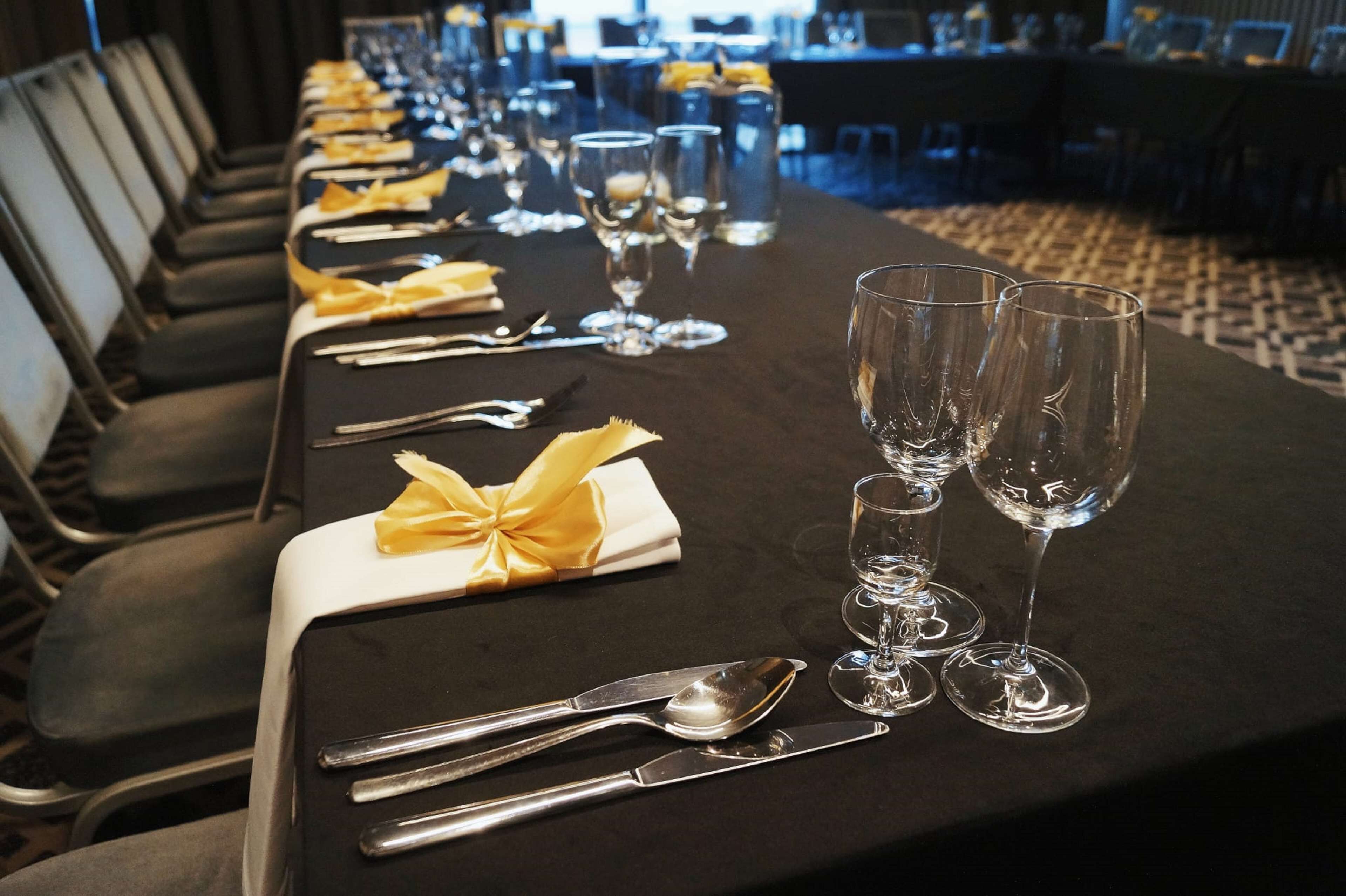 A long banquet table with neatly set dining ware and glasses, chairs aligned on one side, and a patterned carpet.