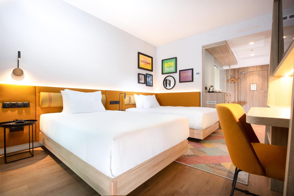Une chambre d’hôtel avec deux lits, un bureau, une chaise et un miroir. Des tableaux encadrés ornent le mur et une lampe se trouve sur la table de chevet.