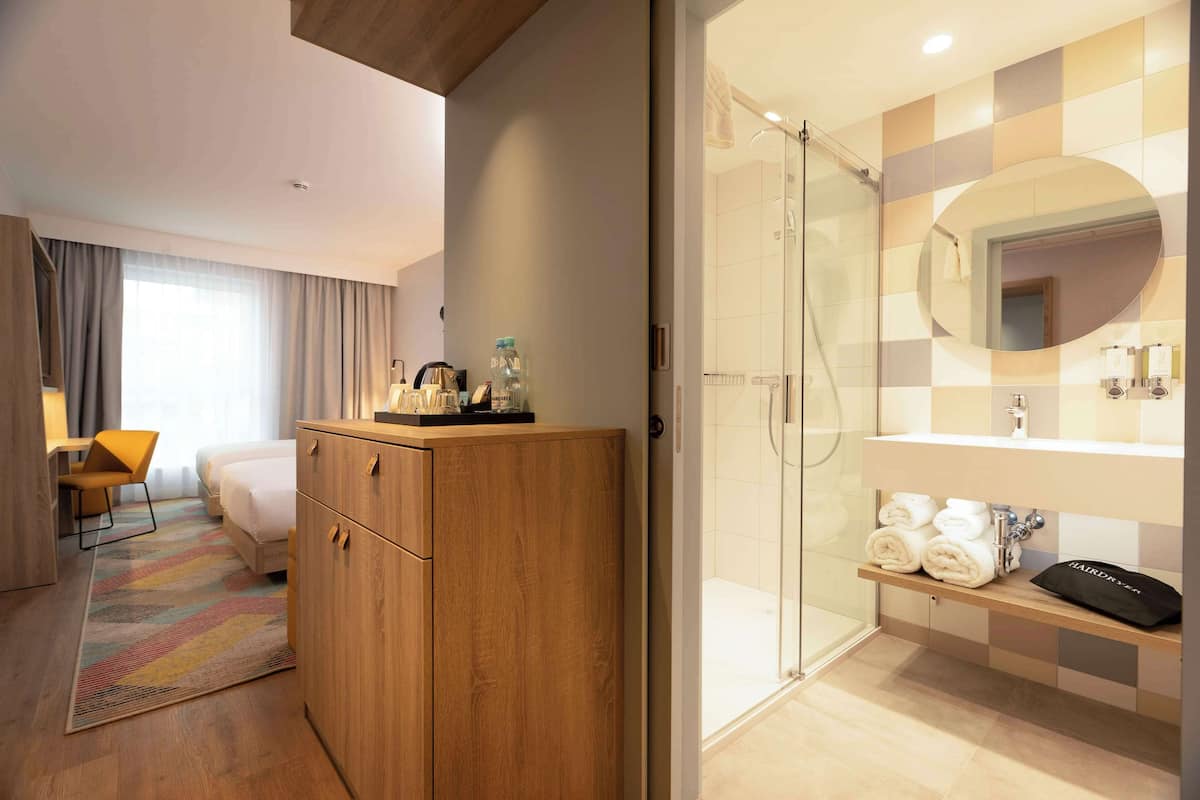 Une chambre d’hôtel comprenant un placard en bois, un lit, un bureau avec une chaise, un espace douche avec une porte en verre, et une salle de bain avec un miroir et des serviettes.