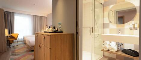 A hotel room with a wooden wardrobe, a bed, a desk with a chair, a shower area with a glass door, and a bathroom with a mirror and towels.