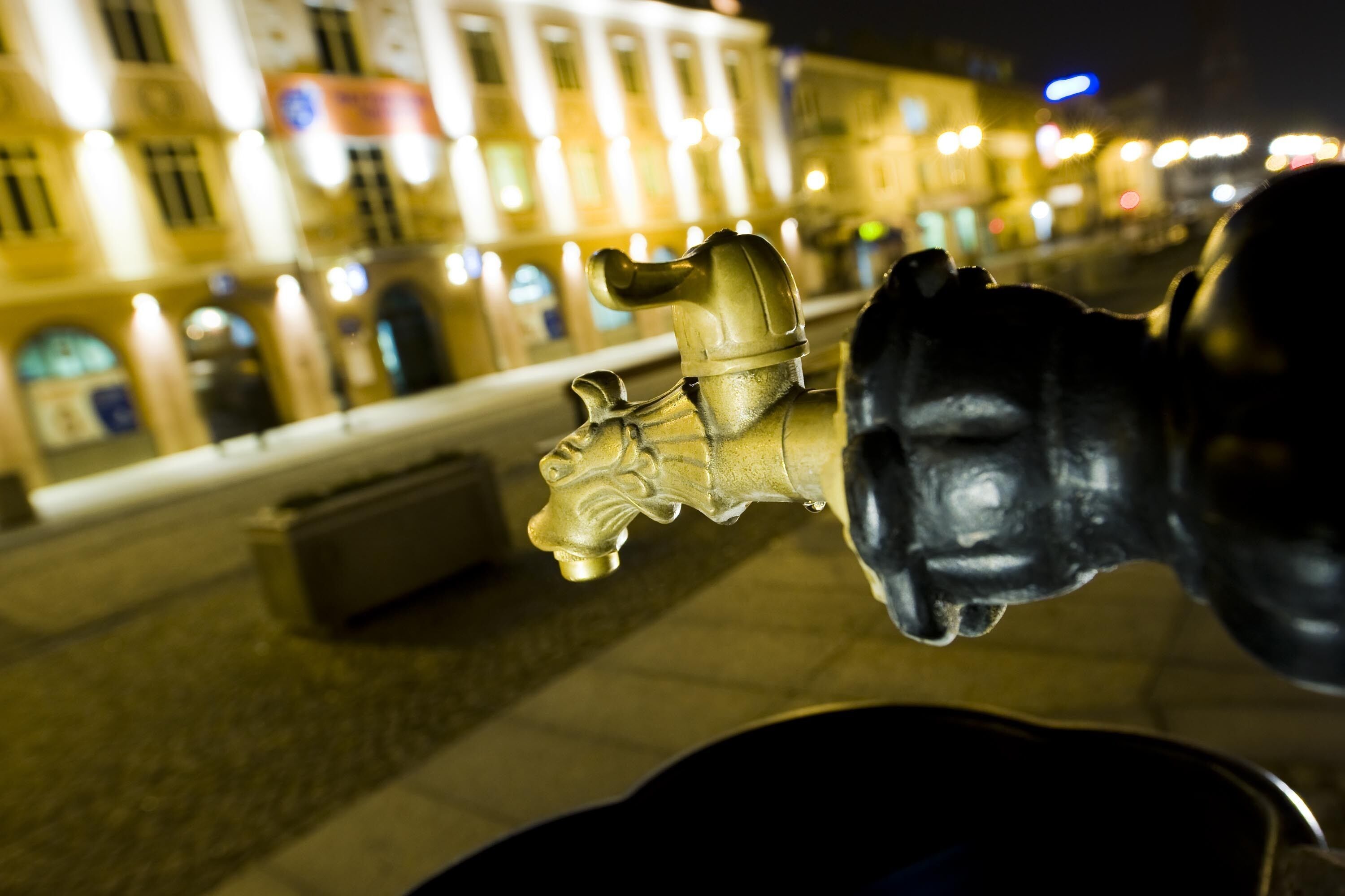 A close-up of a fountain with a lion head spout, set against a backdrop of illuminated buildings at night.