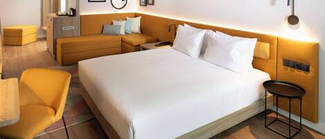 Habitación de hotel con una cama grande, un sofá, una mesita y una silla. Se ve un armario y un espejo en la pared.