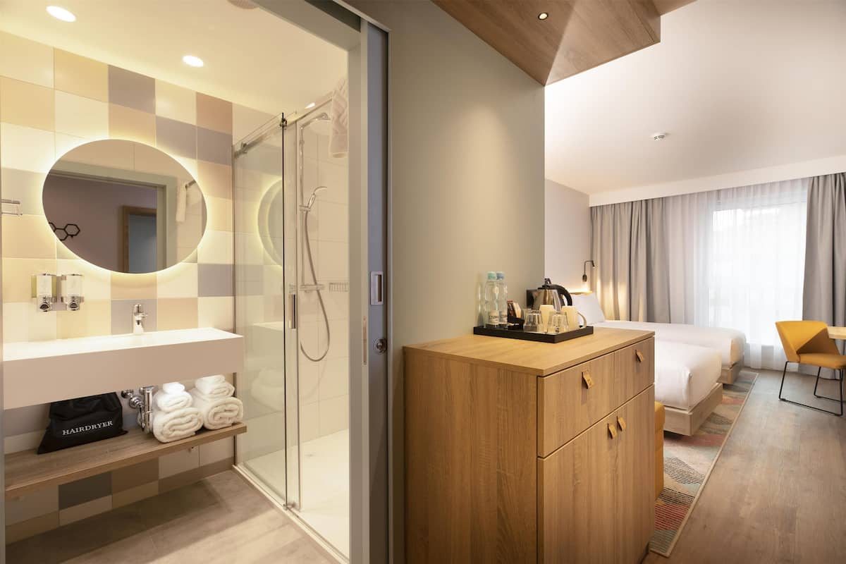 Une chambre d’hôtel moderne équipée d’une douche, d’un grand miroir, d’une commode en bois, d’un lit et d’un bureau avec une chaise.