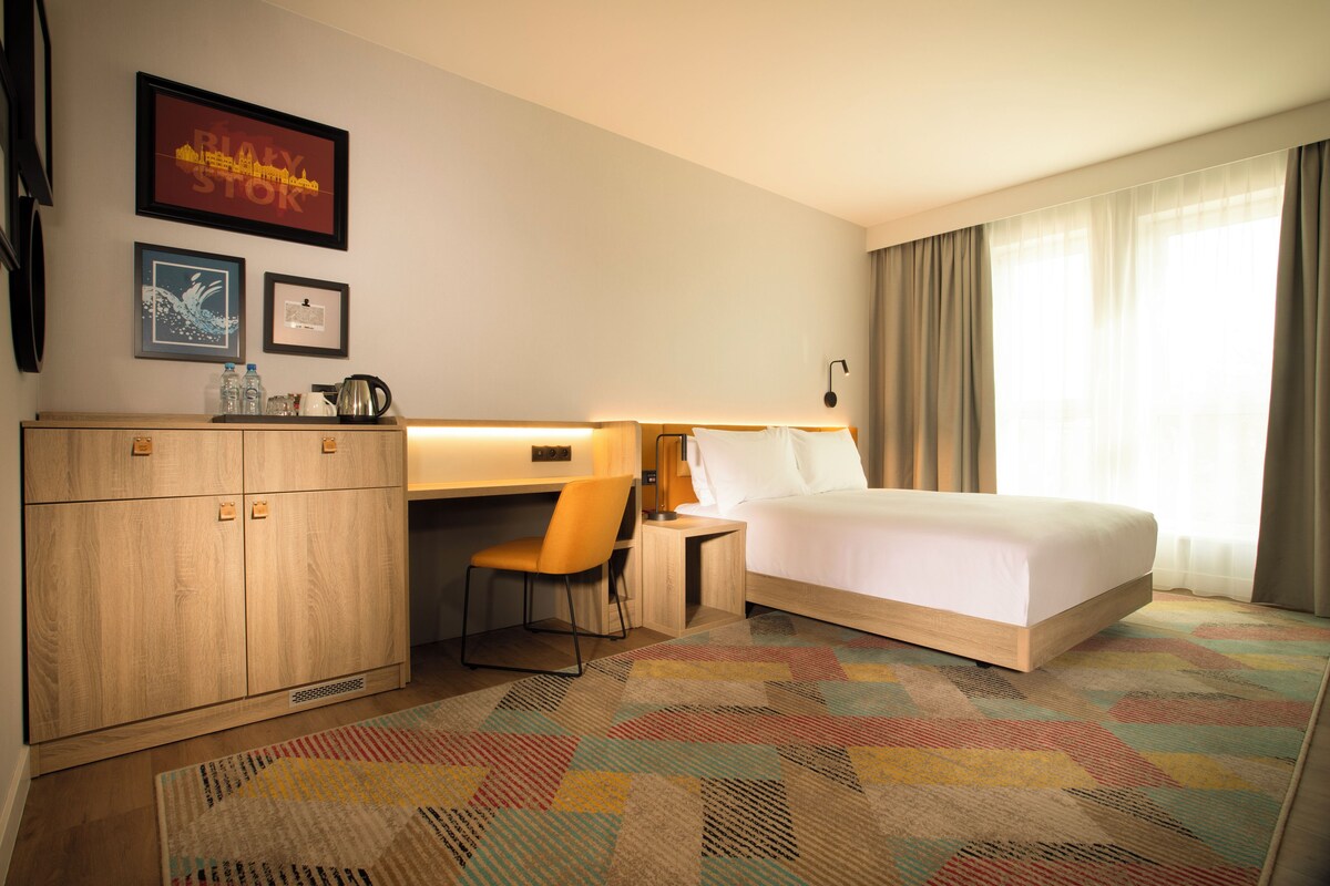 Habitación de hotel con cama, escritorio, silla y un ventanal con cortinas.