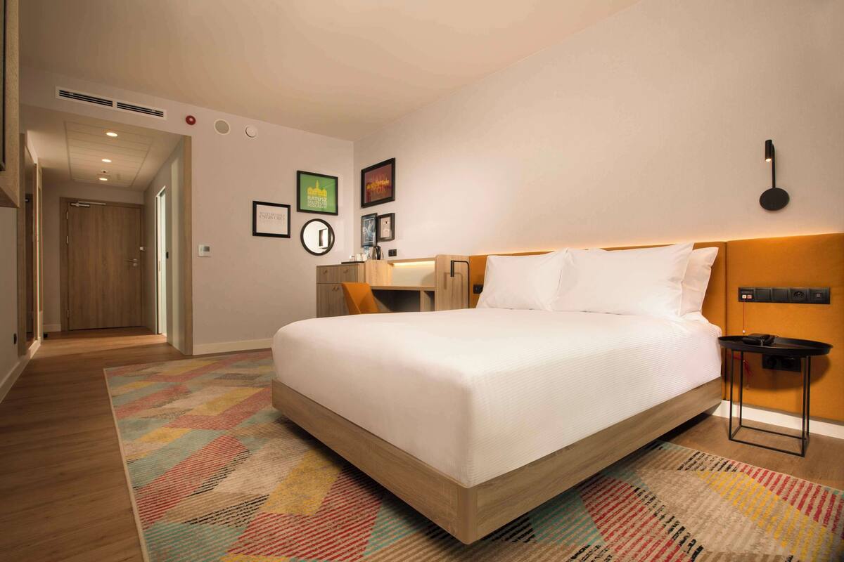 Une chambre d’hôtel avec un grand lit, un bureau et une chaise. Des lampes sont fixées au mur, et une porte donne accès à une autre pièce.