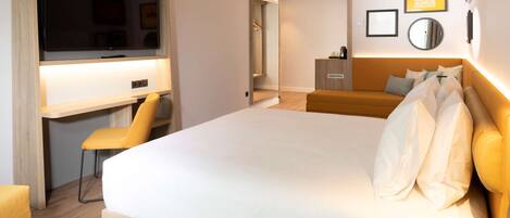 A modern hotel room with a large bed, a desk with a chair, a mirror, and a small kitchenette.