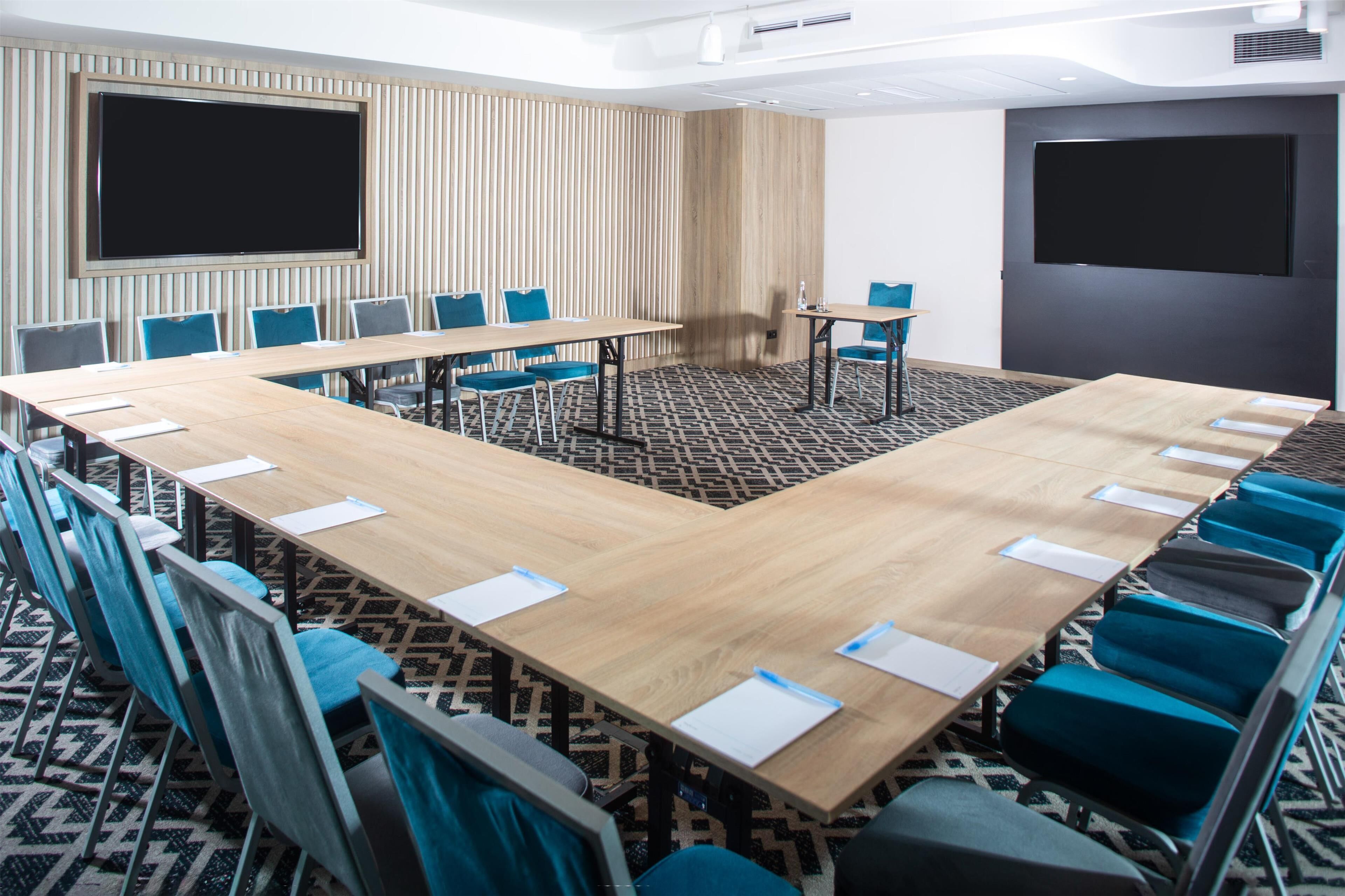 A conference room with a large table, blue chairs, and two screens on the walls.