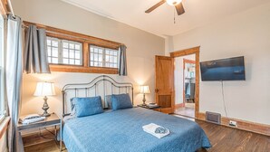 Ein Schlafzimmer mit einem großen Bett, zwei Nachttischen mit Lampen, einem Deckenventilator, einem an der Wand montierten Fernseher und einem Fenster mit Vorhängen.