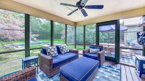 A covered patio with wicker furniture and a ceiling fan, overlooking a backyard with a pool and greenery. - Oasis heaven close to everything you need! (East Hampton)