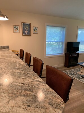 A kitchen with a granite countertop, wooden chairs, a dining table, and a TV stand with a flat-screen TV.