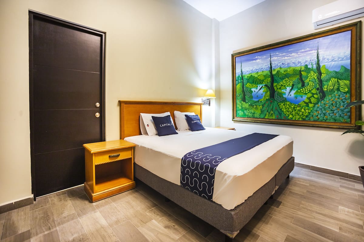 Una habitación de hotel con cama, mesita de noche, un cuadro en la pared y una puerta.