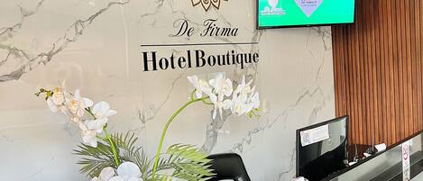 A hotel reception desk with a marble wall, a logo, a television, and a floral arrangement.