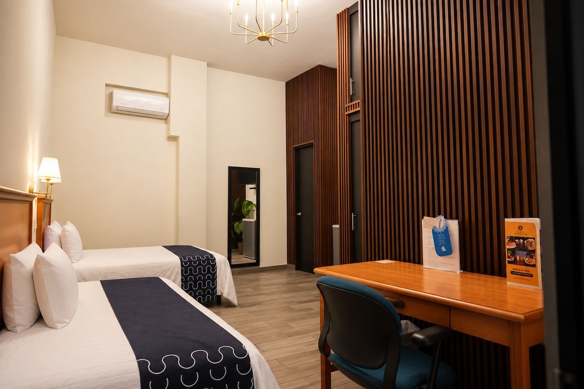A hotel room with two beds, a wooden desk, a chair, a mirror, and a wall-mounted air conditioner.