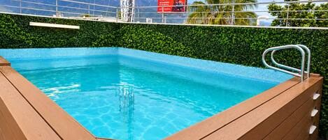 A rectangular swimming pool with a wooden deck and metal handrail, surrounded by a metal fence and a green hedge.