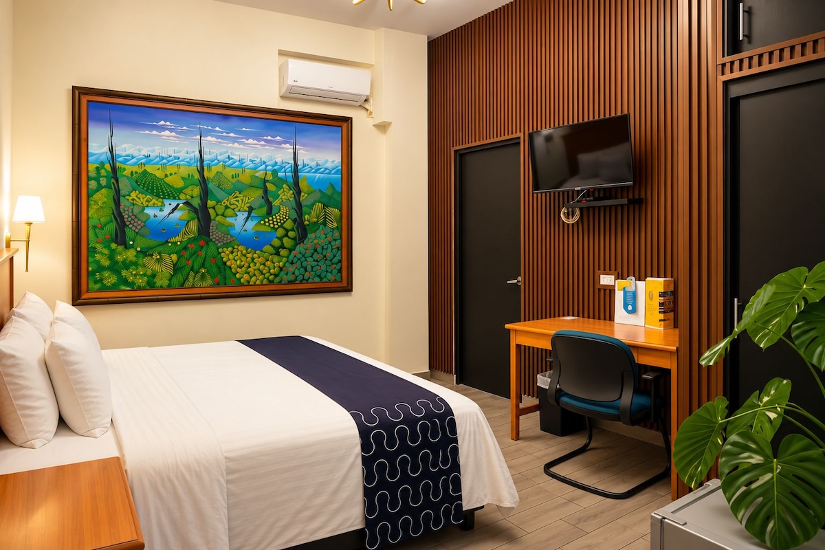 A hotel room with a bed, a desk, a television, and a large landscape painting on the wall.