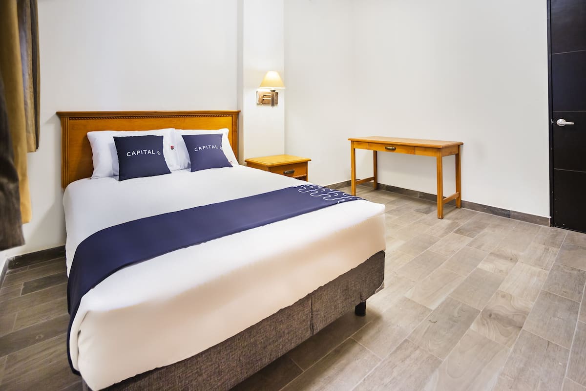 Una cama bien tendida con una colcha azul oscuro y almohadas, una mesita de noche de madera, una lámpara fijada a la pared y una puerta de madera.