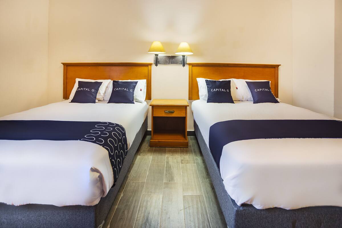Habitación de hotel con dos camas, cada una con almohadas a juego y cabeceros de madera. Un mesita de noche entre las camas con dos lámparas fijadas a la pared.