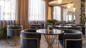 Un moderno vestíbulo de hotel con mesas redondas y sofás, ventanas amplias con cortinas y un espejo grande que refleja el espacio.