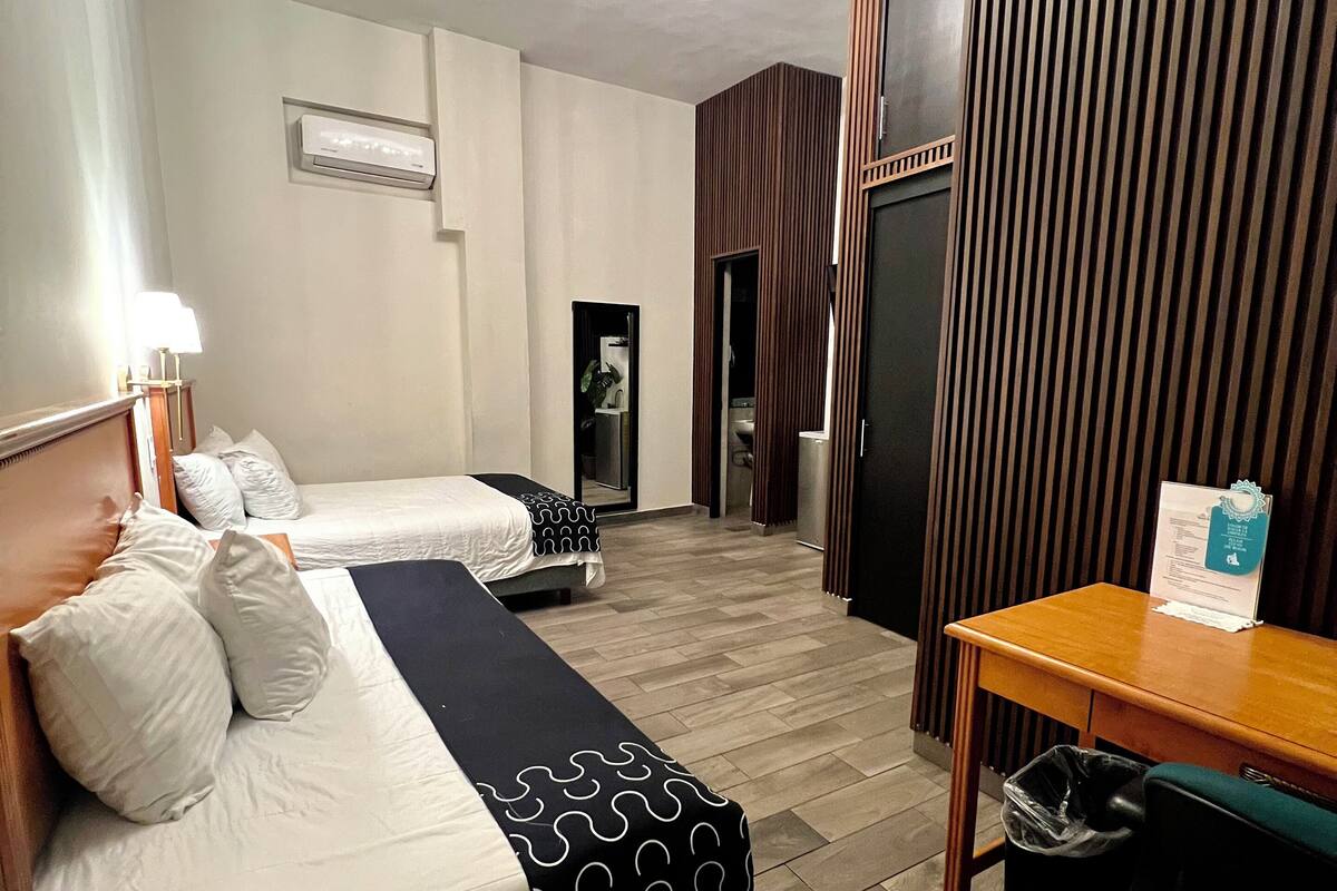 Una habitación de hotel con dos camas, una mesa de madera, un cubo de basura y una araña.