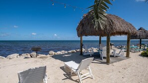 A beach with white lounge chairs, a thatched-roof gazebo, and a wooden deck overlooking the sea. - Free Kayaks Overlooking Pickleball Courts (Key Largo)