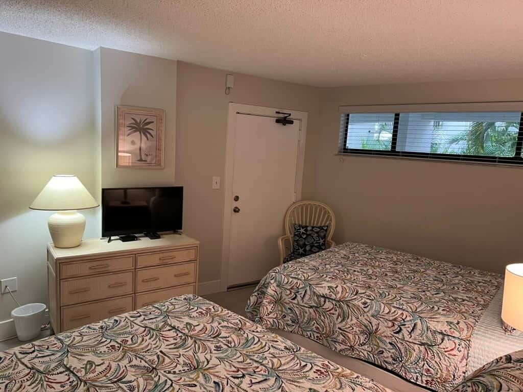 Ein Zimmer mit zwei Betten, einem Fernseher, einem Sessel und einem Fenster.