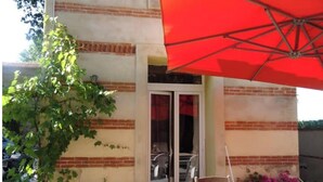 Un espace patio avec un parasol rouge, une table en bois, une plante en pot et une porte donnant sur un espace intérieur.