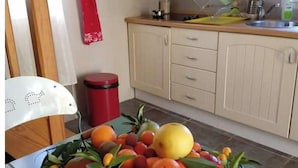 Une cuisine avec un panier de fruits sur la table, une poubelle rouge et une chaise blanche.