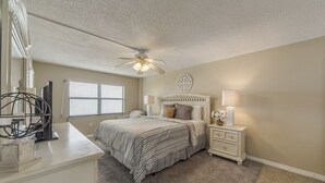 A bedroom with a bed, nightstands, a ceiling fan, a window with blinds, and a decorative wall piece.