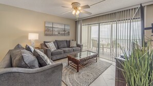 A living room with a grey sofa, a coffee table, and a ceiling fan. There is a large window with blinds and a view of the outdoors.