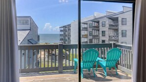 Un balcon avec deux chaises vertes et une terrasse en bois donnant sur une plage et des immeubles d’habitation.