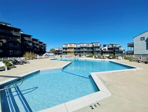 A swimming pool area with a 2ft depth marker, surrounded by modern buildings and lounge chairs.