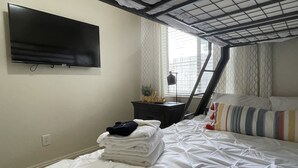 Un dormitorio con cama, televisor montado en la pared, una mesita de noche con lámpara y ventana con cortinas.
