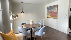 Un salón moderno con una mesa de comedor redonda, sillas azules, un sofá con un cojín color mostaza, una lámpara de pie y un cuadro en la pared.