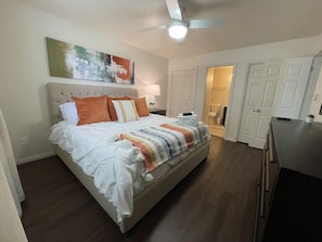 Een slaapkamer met een bed, een plafondventilator, een schilderij en een deur naar de badkamer.