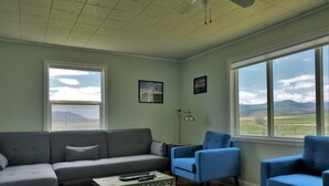 A living room with a grey sofa, a blue armchair, and a patterned ottoman. There are two windows offering a view of the outdoors. - Chesterfield-Lava Hot Springs - Farm Cottage (Bancroft)