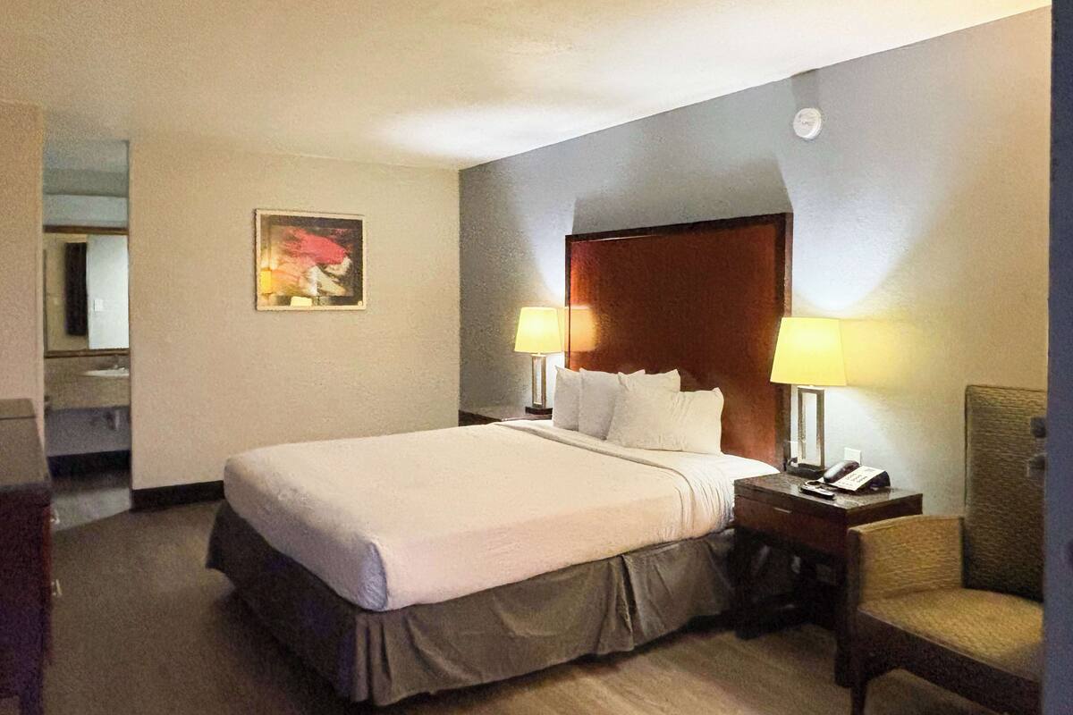 A hotel room with a large bed, two bedside lamps, a chair, a wooden headboard, and a painting on the wall.